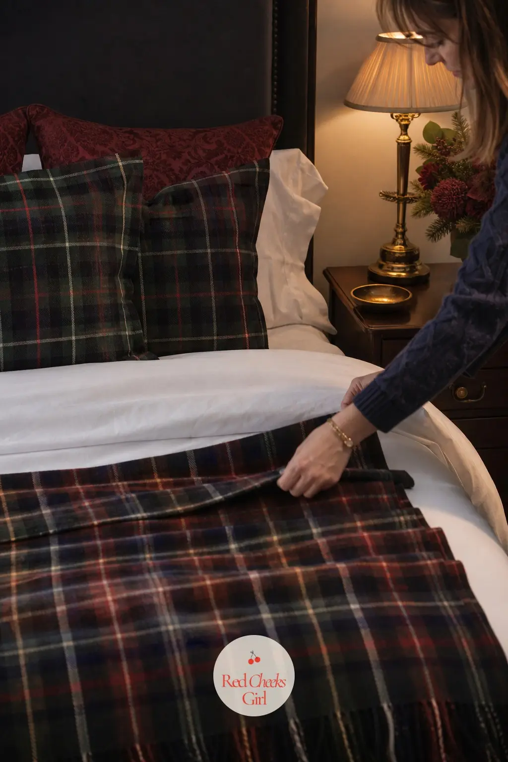 A Guest Room Ready For Overnight Company — A tartan throw lies folded across the foot of the bed, a brass lamp sits lit on the nightstand, and a small dish