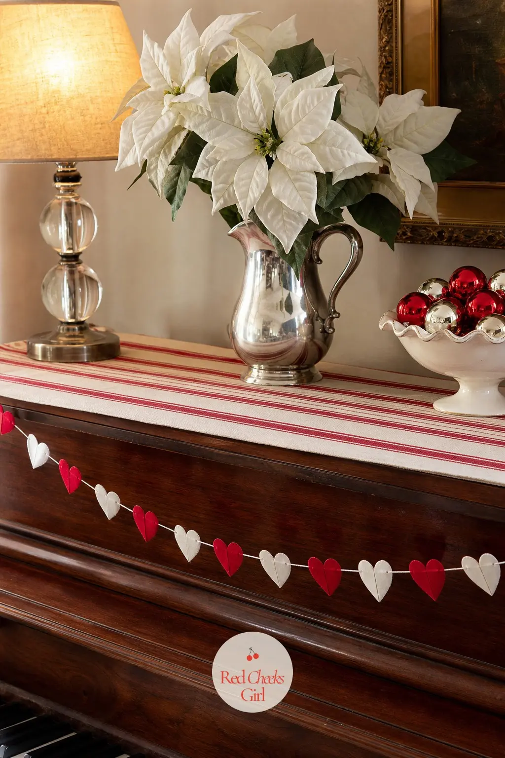 The Piano Top — A silver pitcher holds a handful of white faux poinsettia stems, set on a striped table runner so the piano reads as styled rather than