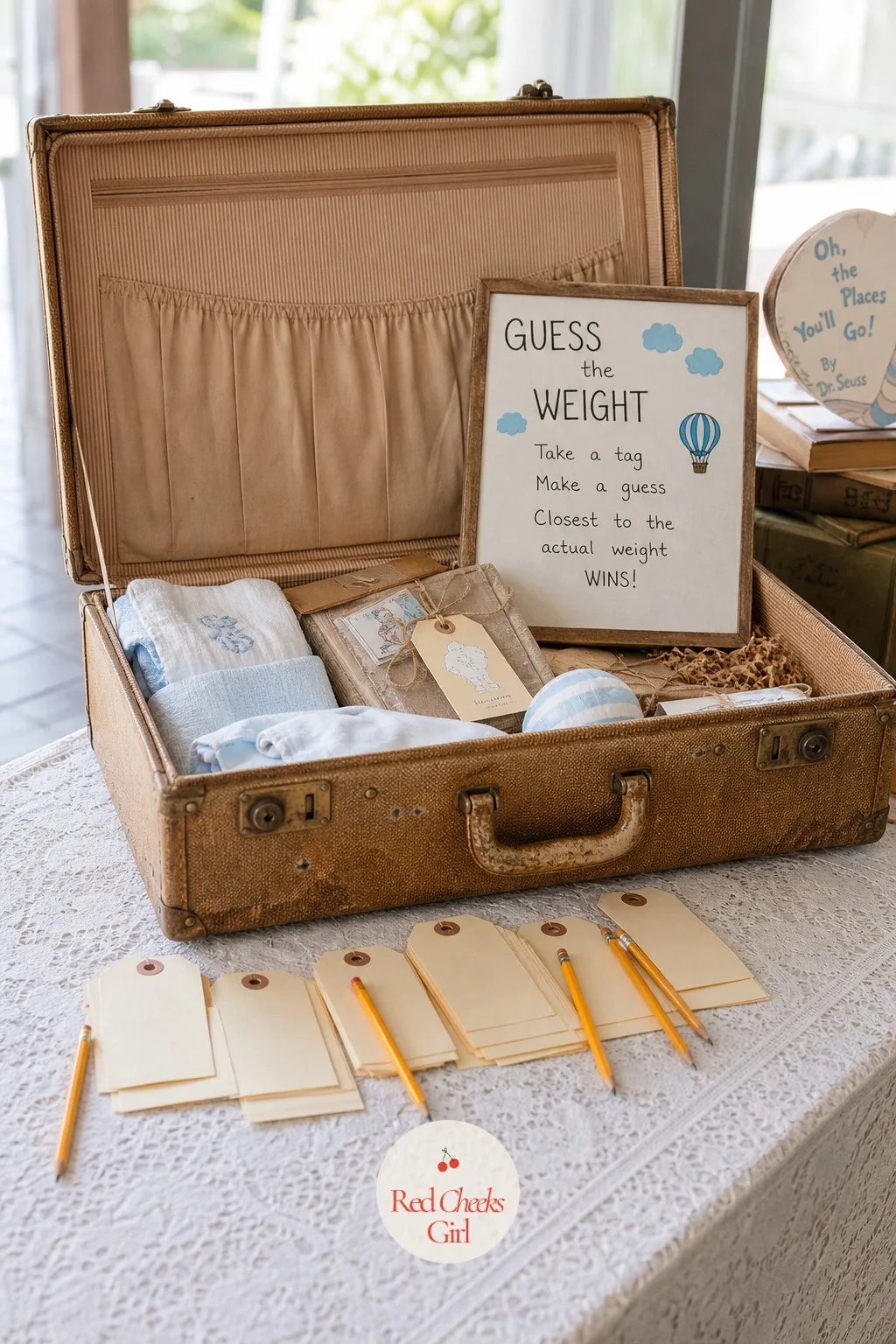 Luggage Tag Guess the Weight Game — A worn suitcase lies open on the gift table with a pile of blank manila luggage tags and a handful of pencils scattered