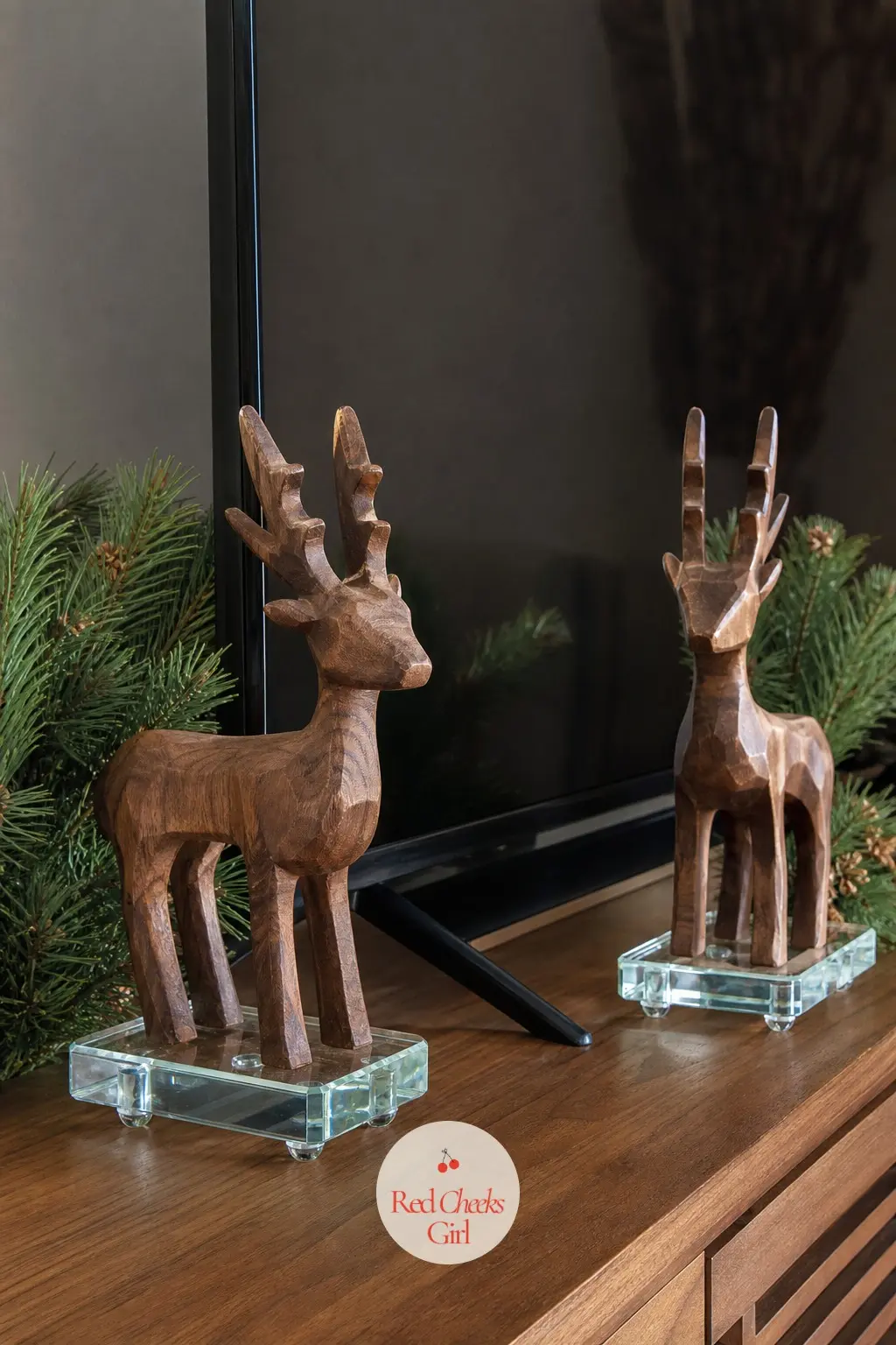 A Pair of Carved Walnut Reindeer Flanking the Screen on Low Glass Risers — Two small walnut-toned reindeer, no taller than a paperback, stand on low glass