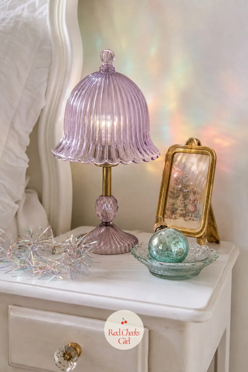 The Nightstand — A small lamp with a lavender glass shade sits beside the bed, its light already doing most of the work, with one tiny glass bauble in aqua
