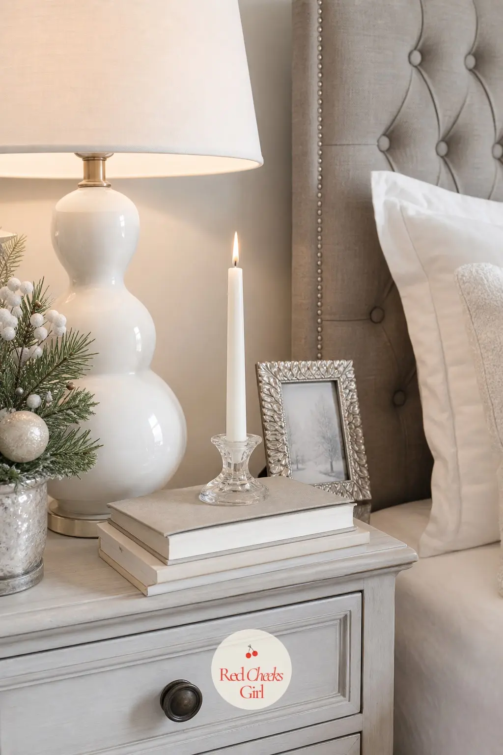 The Bedside Table, Lit for Reading — A ceramic lamp in plain white sits beside a short stack of pale hardcover books and a single taper candle, unlit by day