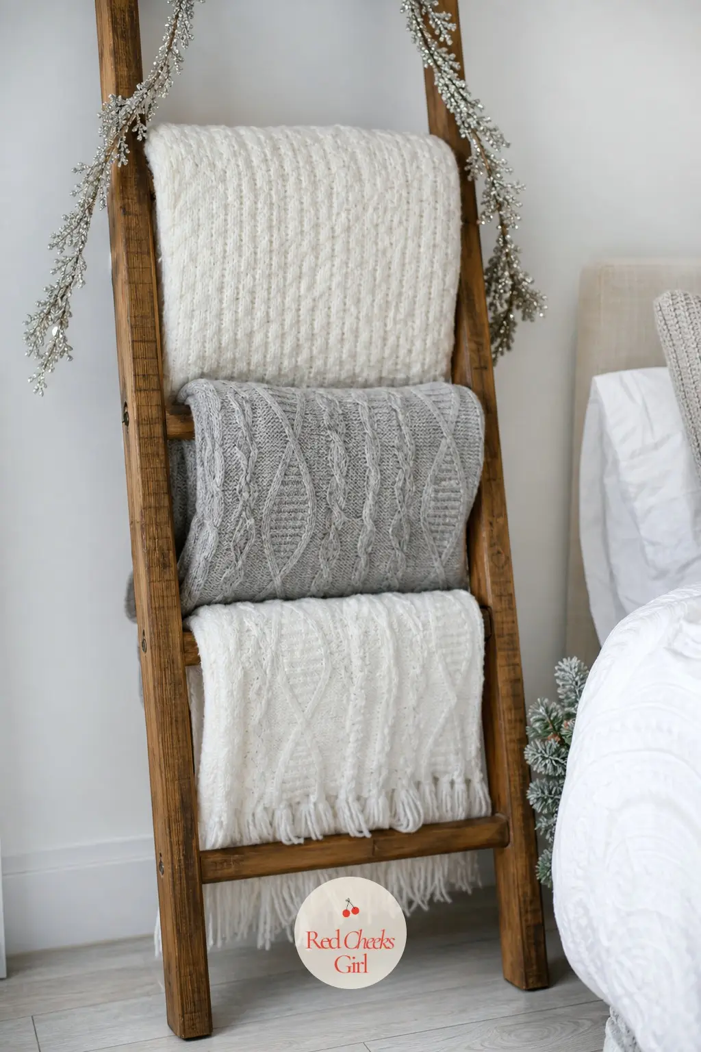 The Blanket Ladder, Stacked in White Throws — A wooden ladder leaned against the wall holds three knit throws in white and dove grey, each one folded to a