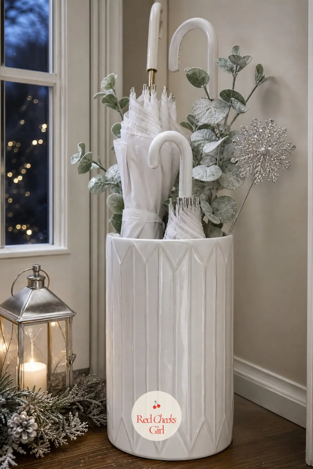 The Umbrella Stand, Topped With a Silver Pick — A plain white ceramic stand holds umbrellas on one side and a bundle of frosted eucalyptus on the other, tied
